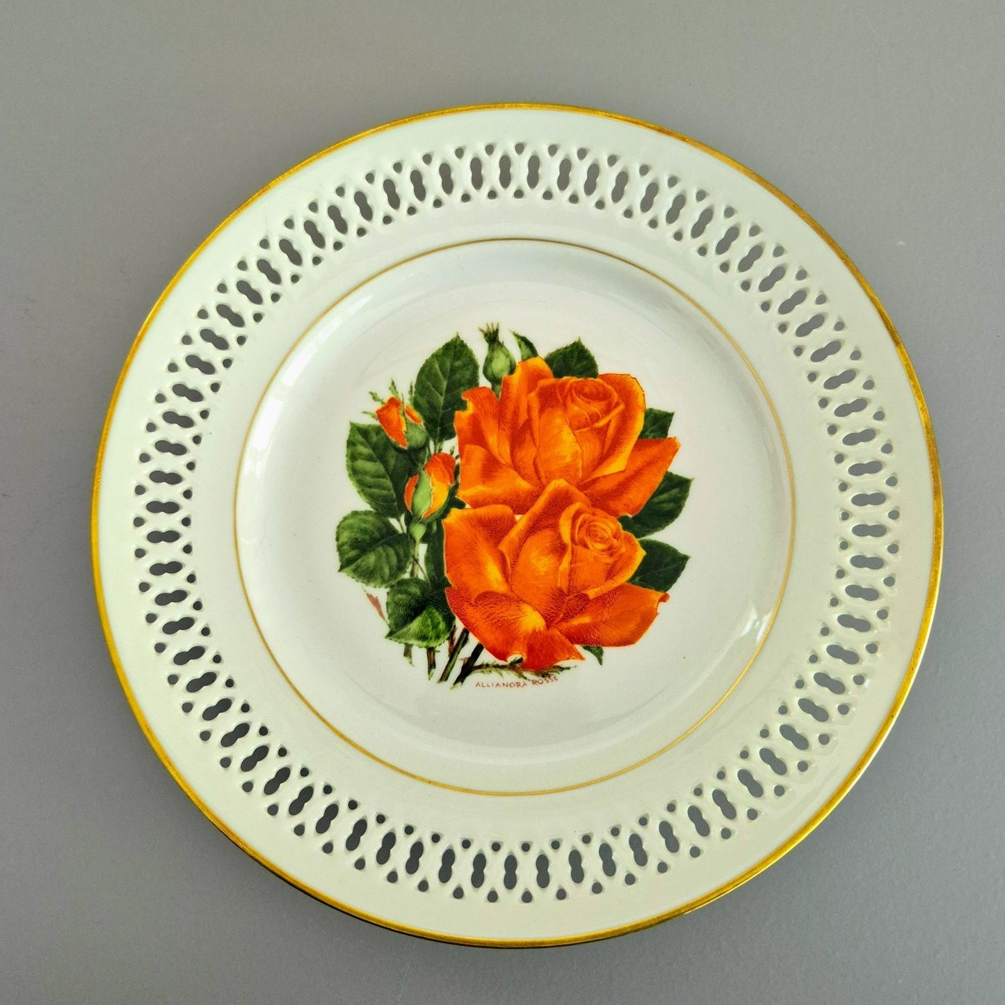 A round white porcelain plate with a gold rim, featuring a decorative border of perforated cutouts and a central illustration of an orange rose with green leaves.