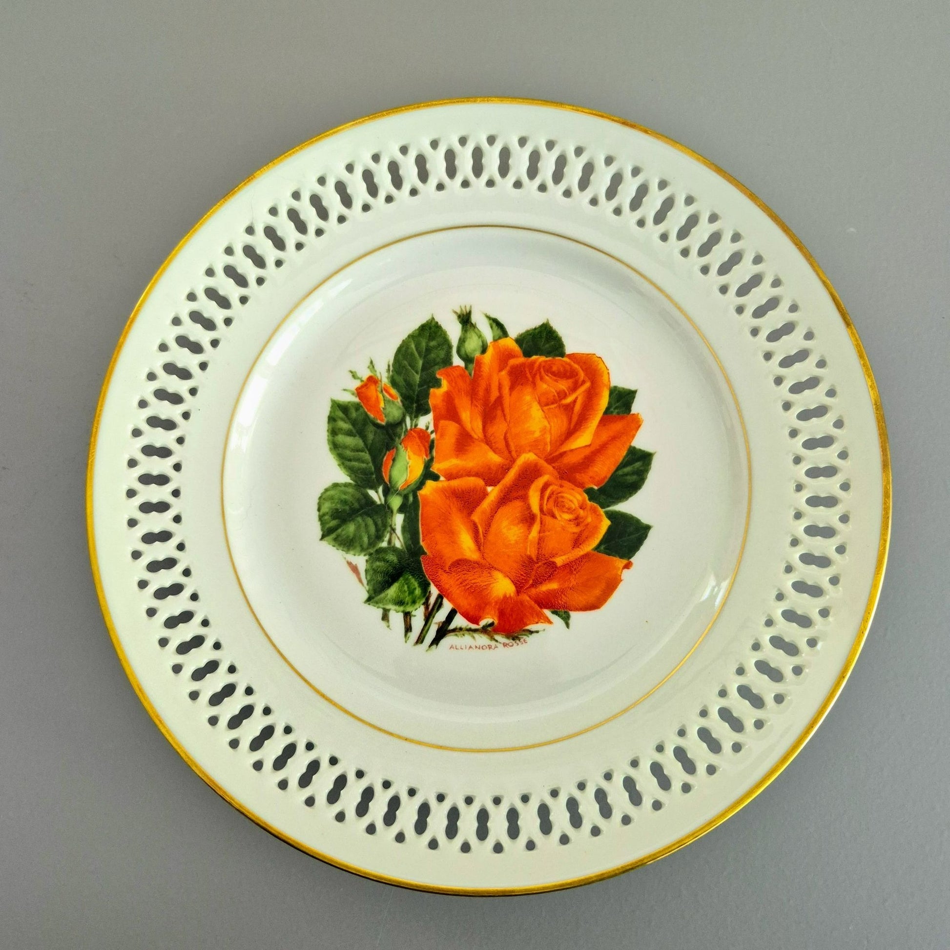 A round white porcelain plate with a gold rim, featuring a decorative border of perforated cutouts and a central illustration of an orange rose with green leaves.