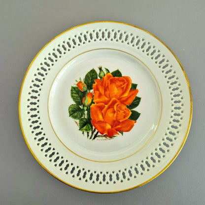 A round white porcelain plate with a gold rim, featuring a decorative border of perforated cutouts and a central illustration of an orange rose with green leaves.